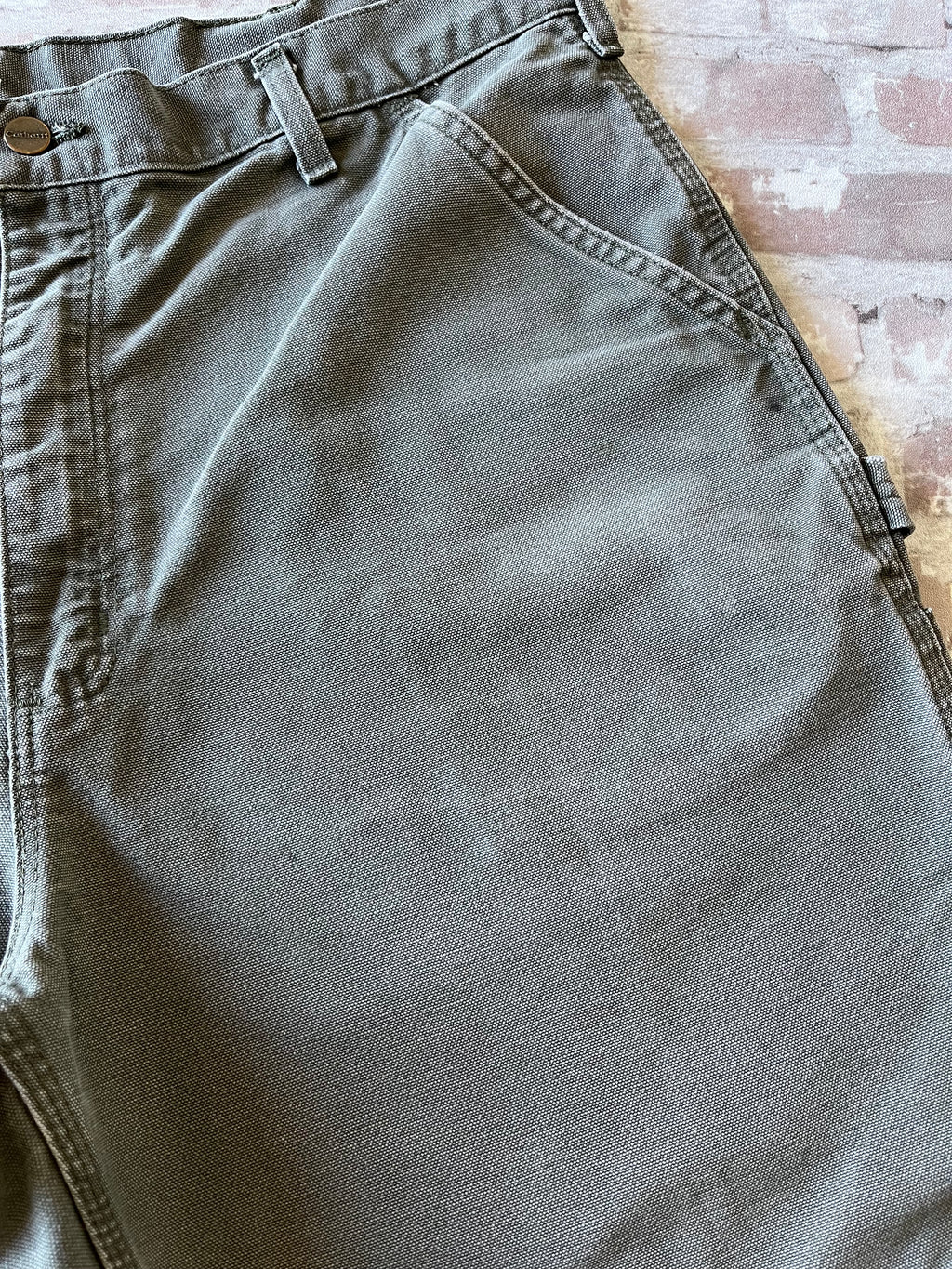 Bermuda de travail vert Moss Carhartt B25 à plat, mettant en valeur la toile en coton canvas délavée et la boucle marteau, focus taches ultra discrètes