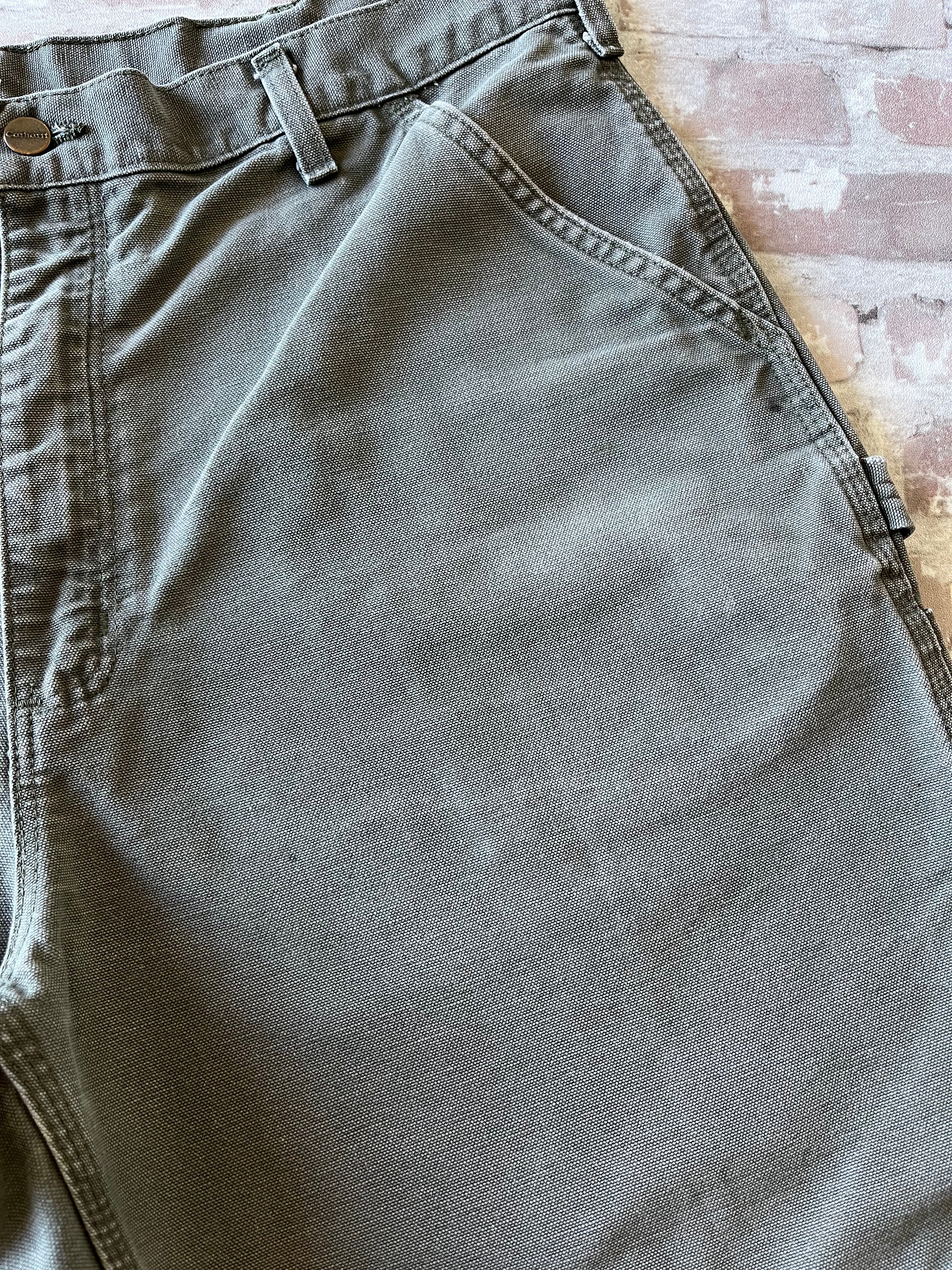 Bermuda de travail vert Moss Carhartt B25 à plat, mettant en valeur la toile en coton canvas délavée et la boucle marteau, focus taches ultra discrètes
