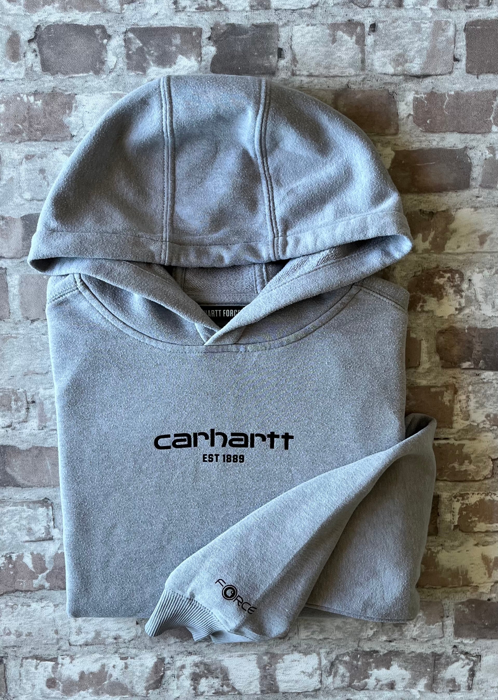 Sweat à capuche vintage Carhartt Force gris clair chiné avec imprimé EST 1889 sur la poitrine et écusson tissé sur la poche, présenté plié à plat.
