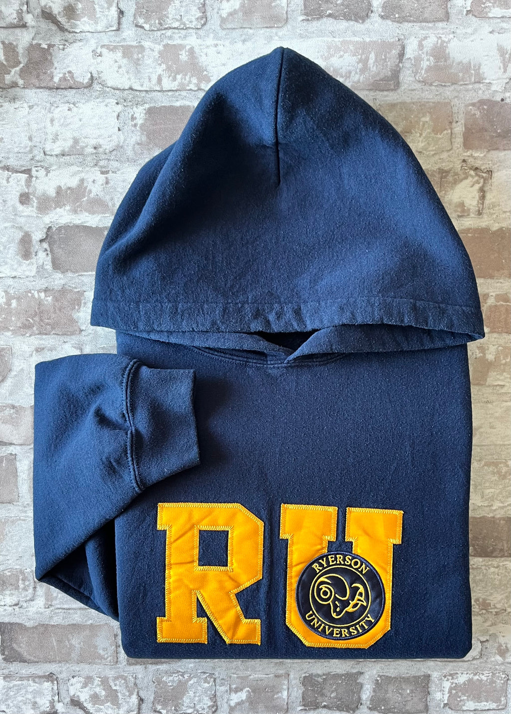 Sweat à capuche vintage bleu marine Ryerson Rams avec écusson bélier brodé et grand lettrage RU en satin jaune, présenté plié à plat.