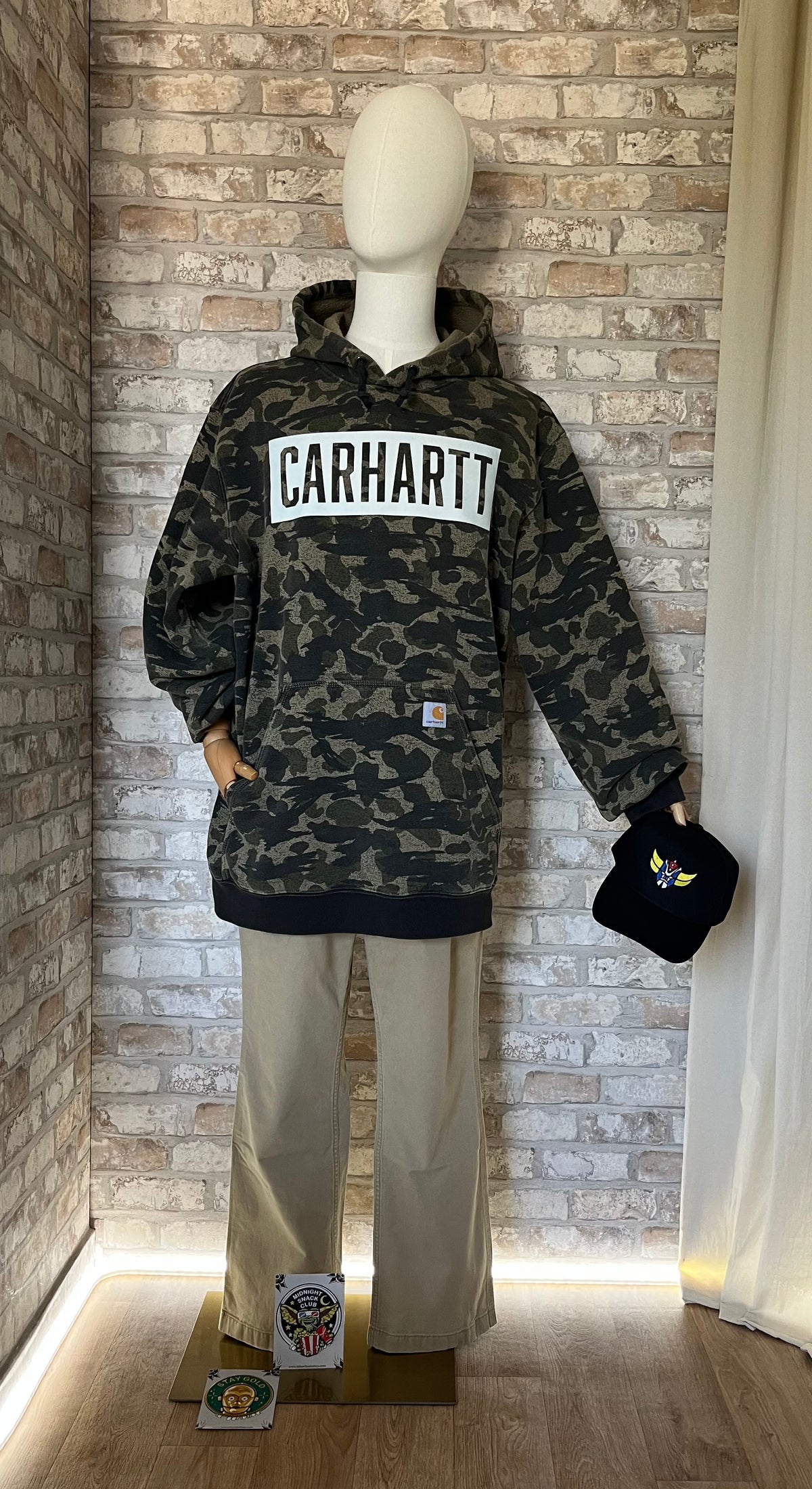 Hoodie Big Logo Carhartt coloris Kaki Beige Camouflage collection Fall-Winter, porté sur un mannequin de face avec un pantalon Carhartt, une casquette Goldorak Grafitee à la main et des Patchs La Barbuda posés au sol