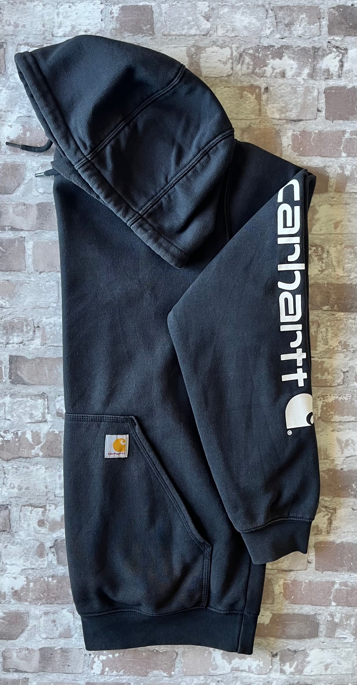 Sweat à capuche noir Carhartt K288 Heavyweight à patine décolorée avec écusson de la marque sur la poche kangourou, présenté à plat, de profil