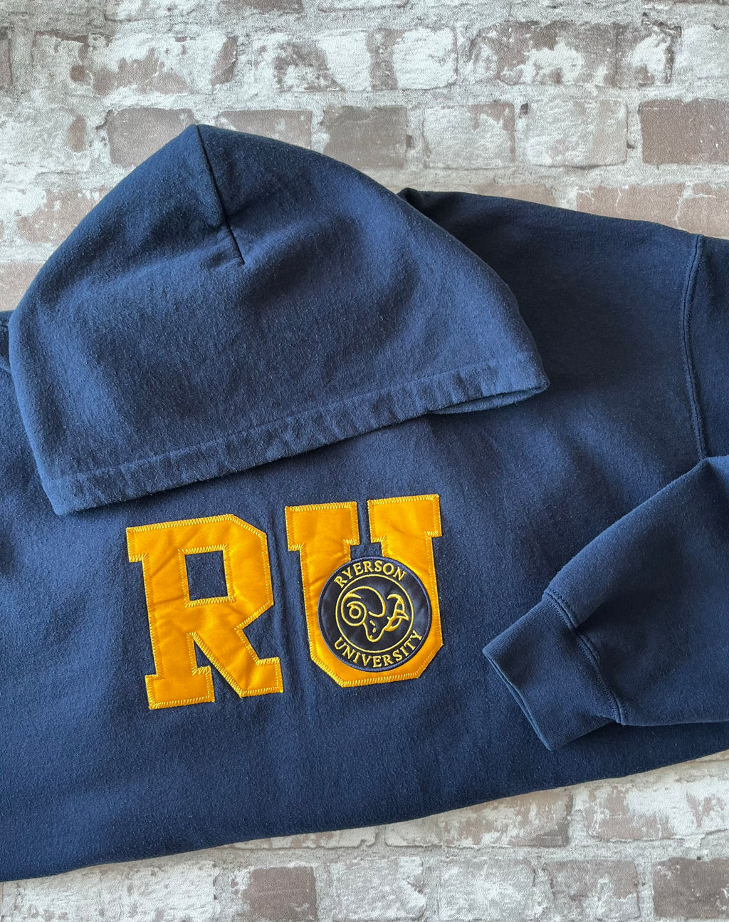 Sweat à capuche vintage bleu marine Ryerson Rams avec écusson bélier brodé et grand lettrage RU en satin jaune, présenté plié à plat.