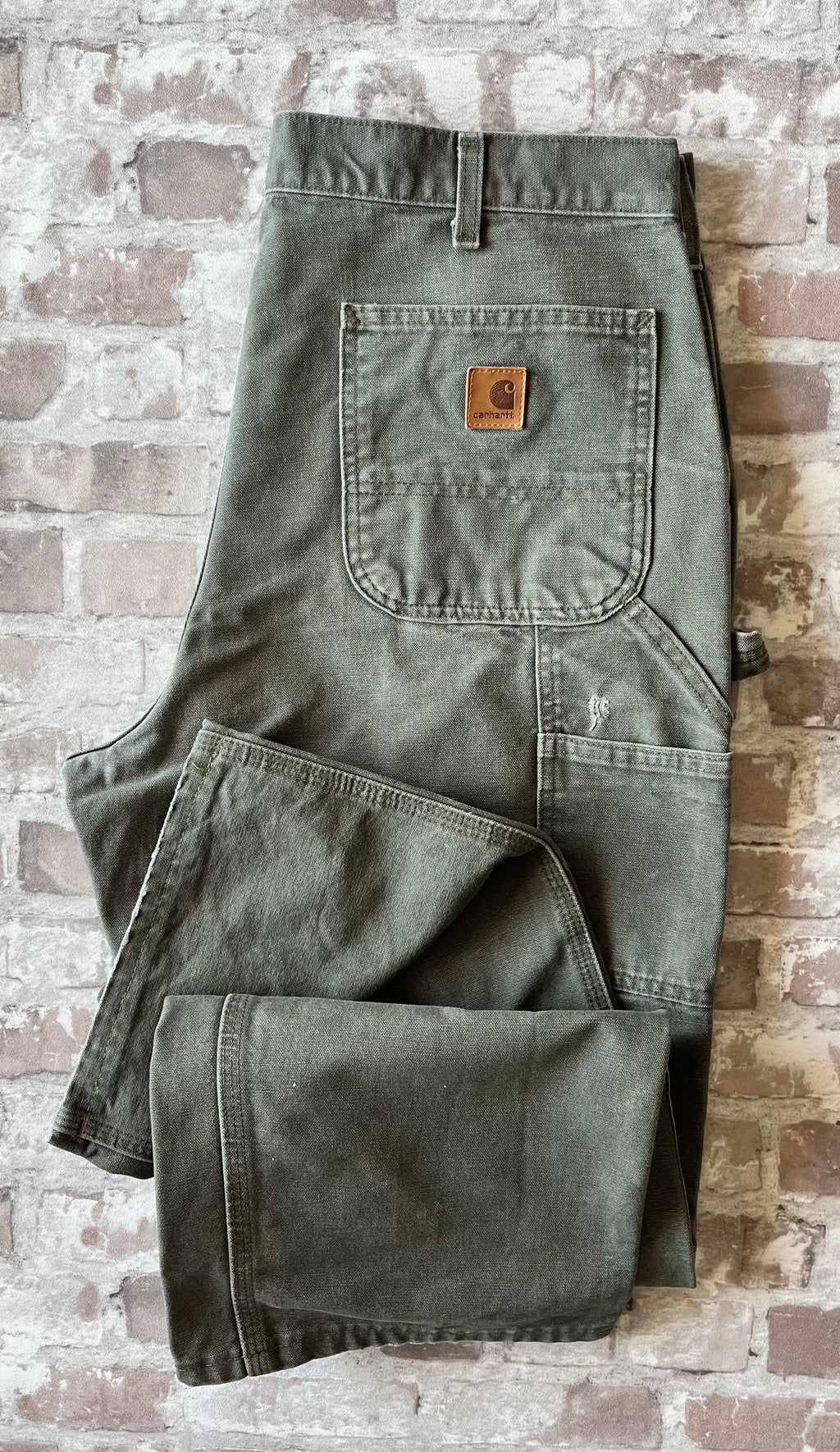 Pantalon de travail vert Moss Carhartt B11 plié à plat, mettant en avant la toile Duck Canvas lourde et la poche arrière avec patch logo ( et un accroc visible )