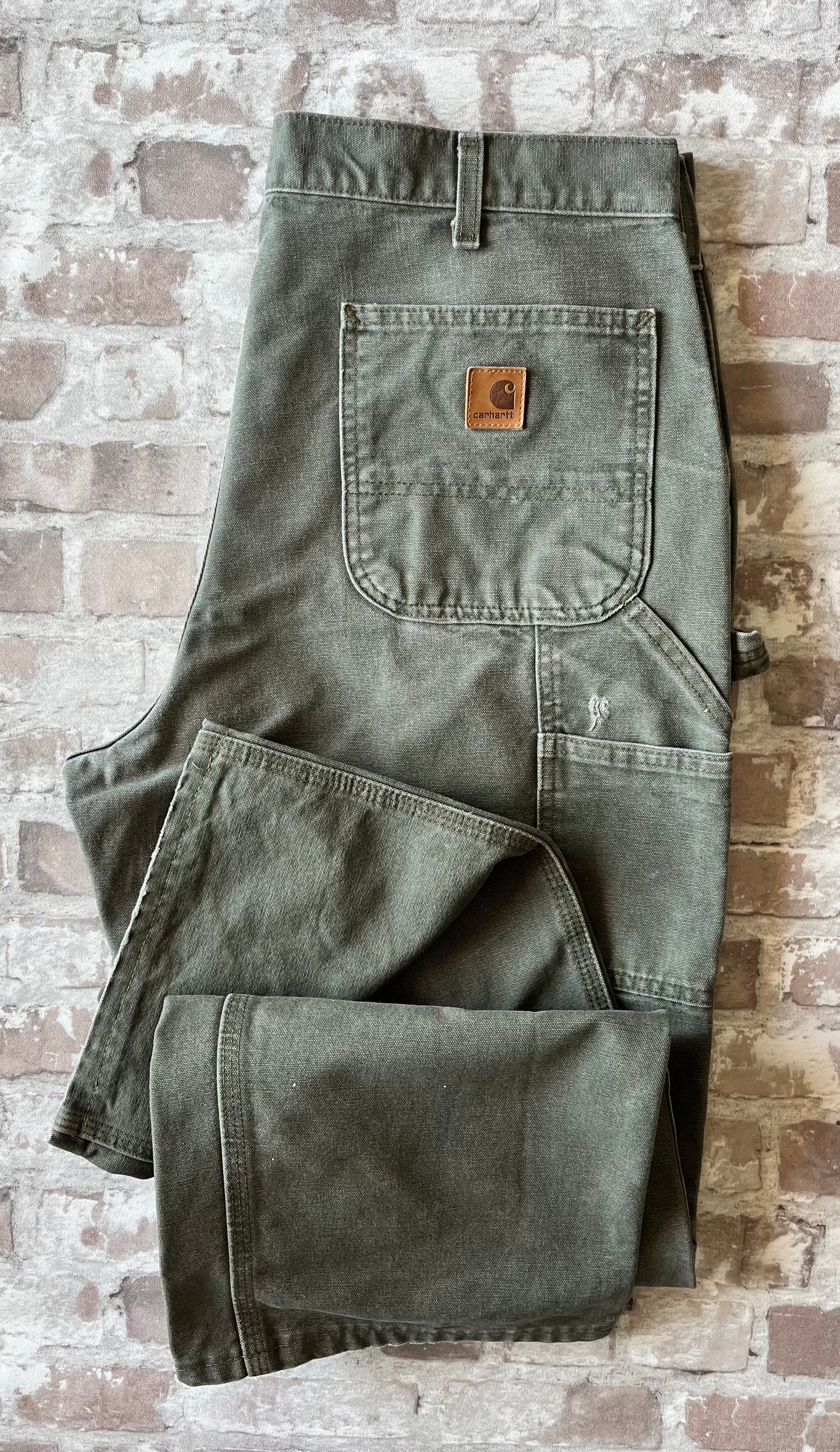 Pantalon de travail vert Moss Carhartt B11 plié à plat, mettant en avant la toile Duck Canvas lourde et la poche arrière avec patch logo ( et un accroc visible )