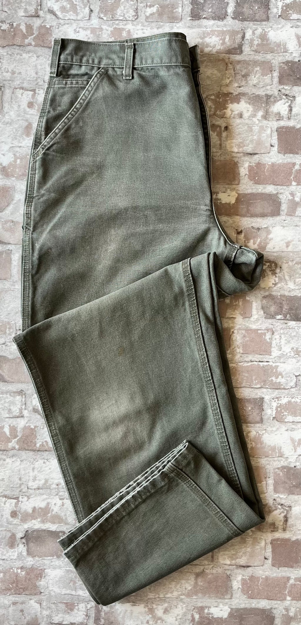 Pantalon de travail vert Moss Carhartt B11 plié à plat, mettant en avant la toile Duck Canvas lourde et la patinée