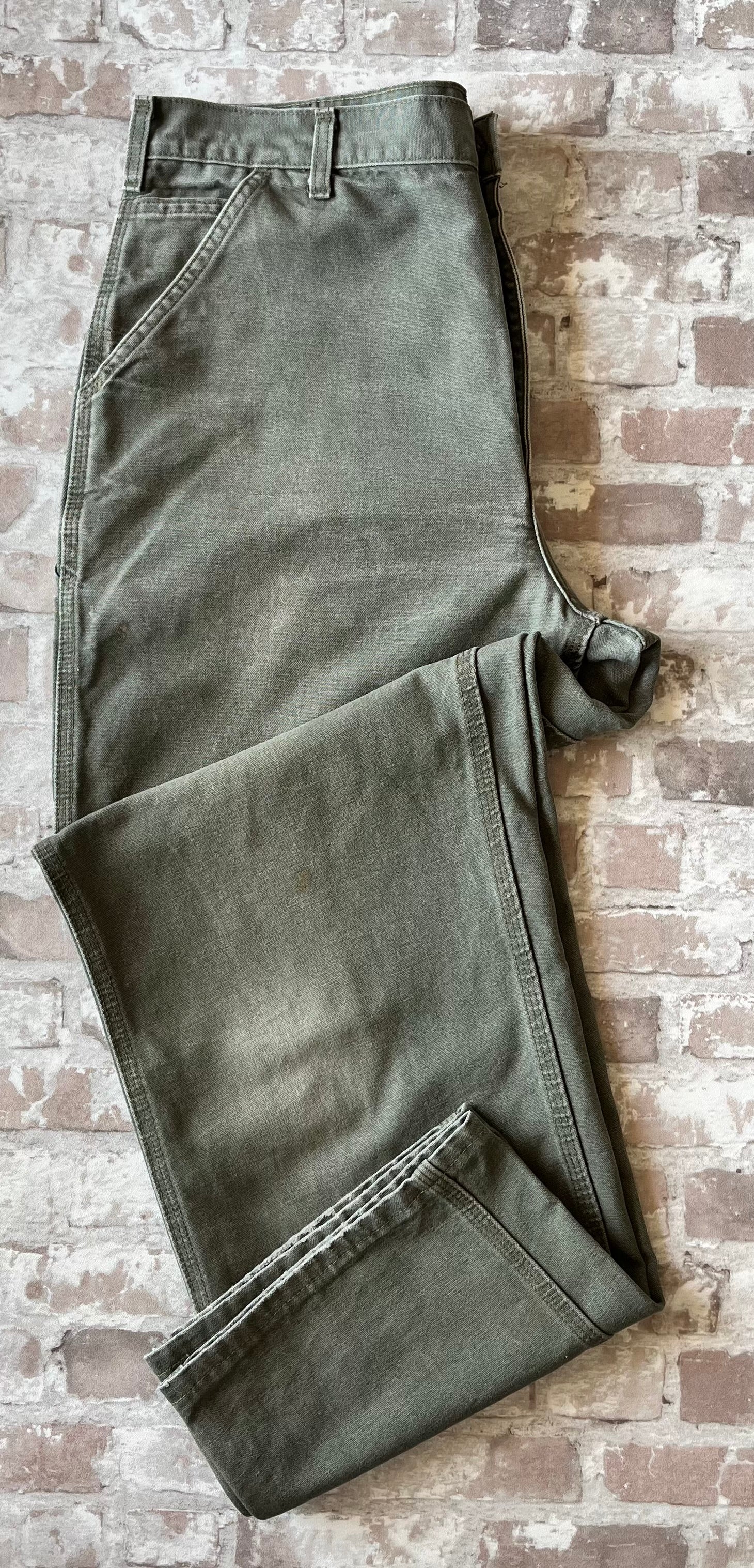 Pantalon de travail vert Moss Carhartt B11 plié à plat, mettant en avant la toile Duck Canvas lourde et la patinée