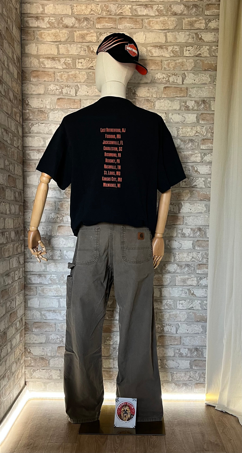 Pantalon Carpenter Carhartt B151, Canvas Dungaree Fit, Light Brown Faded exposé sur mannequin de dos avec un T-shirt Rock/tour Bruce Springsteen, une casquette vintage Harley Davidson et un Patch La Barbuda au sol.