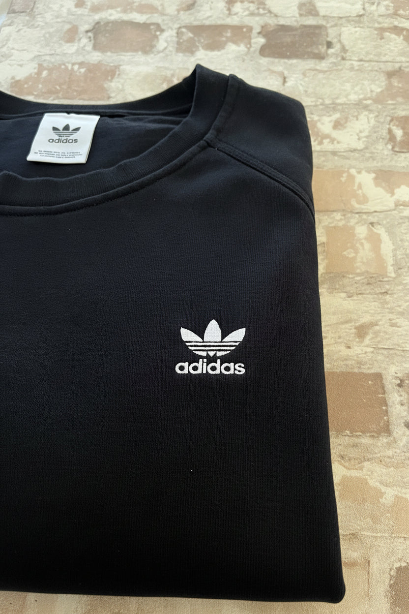Sweat col rond vintage Adidas Originals noir avec l'iconique logo trèfle blanc brodé sur la poitrine, présenté plié à plat, gros plan logo