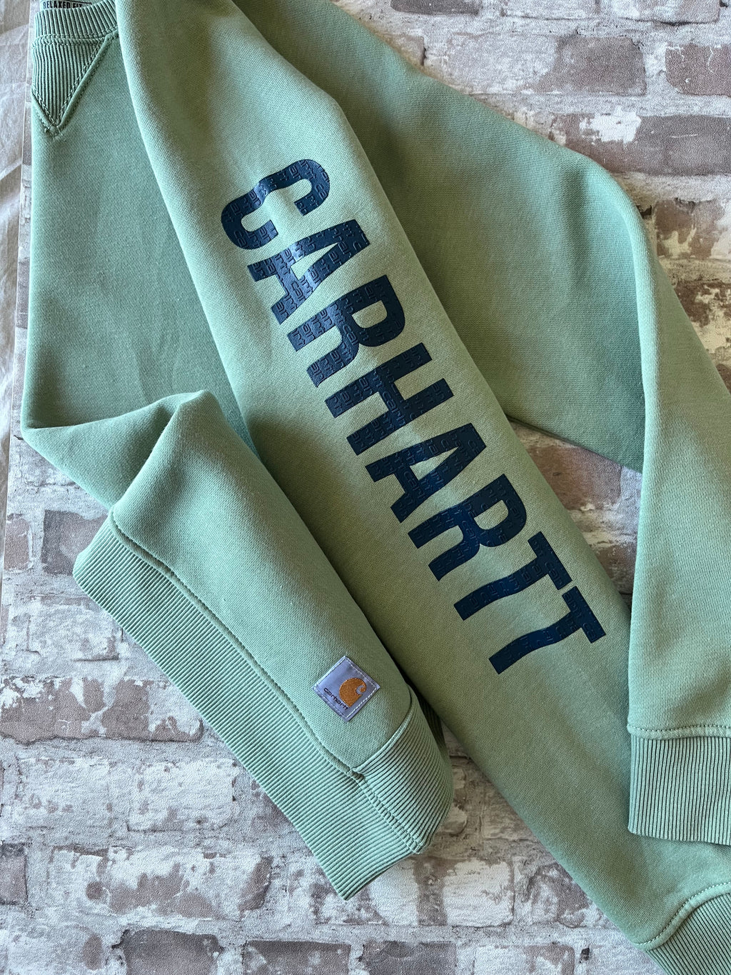 Gros plan sur le logo texturé bleu marine imprimé le long de la manche gauche d'un sweat vert sauge Carhartt.