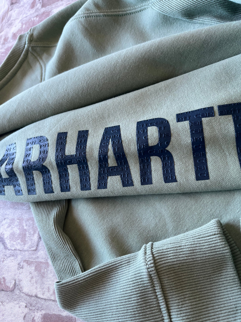 Gros plan sur le logo texturé bleu marine imprimé le long de la manche gauche d'un sweat vert sauge Carhartt.