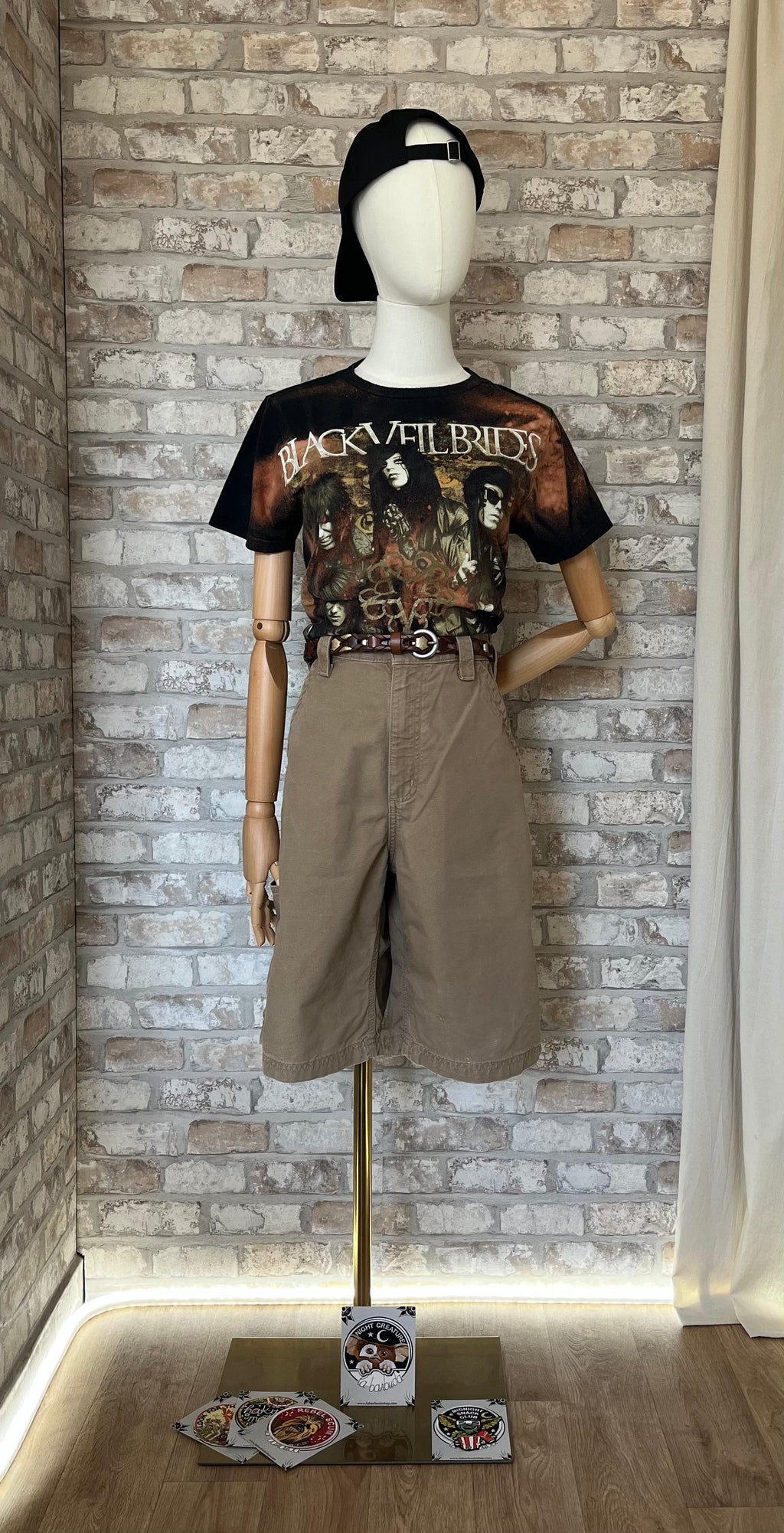 Silhouette streetwear portée par un mannequin avec un t-shirt noir Black Veil Brides, un short carpenter DKH Carhartt, une ceinture en cuir Fossil et une casquette Goldorak. Des Patchs La Barbuda au sol