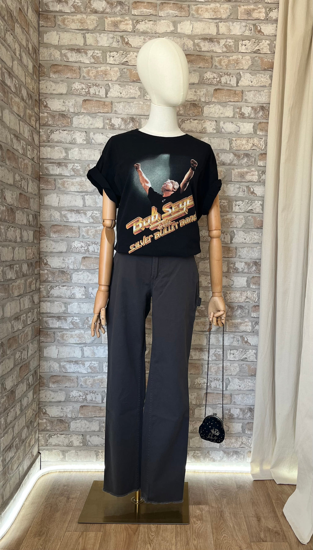 Silhouette femme portée par un mannequin avec un t-shirt noir Bob Seger, un pantalon gris anthracite Carhartt et un sac bourse IKKS, vue de face.
