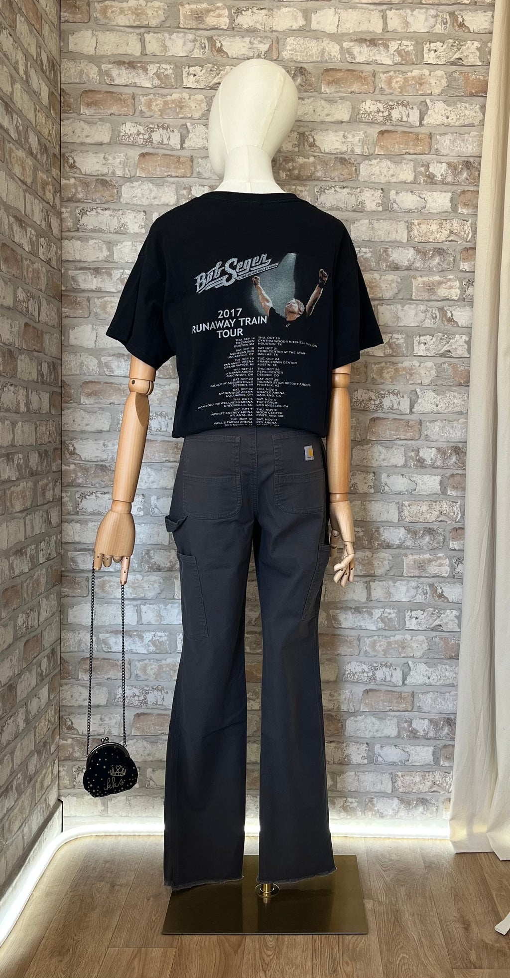 Vue de dos d'un mannequin portant un pantalon de travail gris Carhartt avec patch logo sur la poche arrière, un rock Tee Band Tour Bob Seger et un sac bourse IKKS.