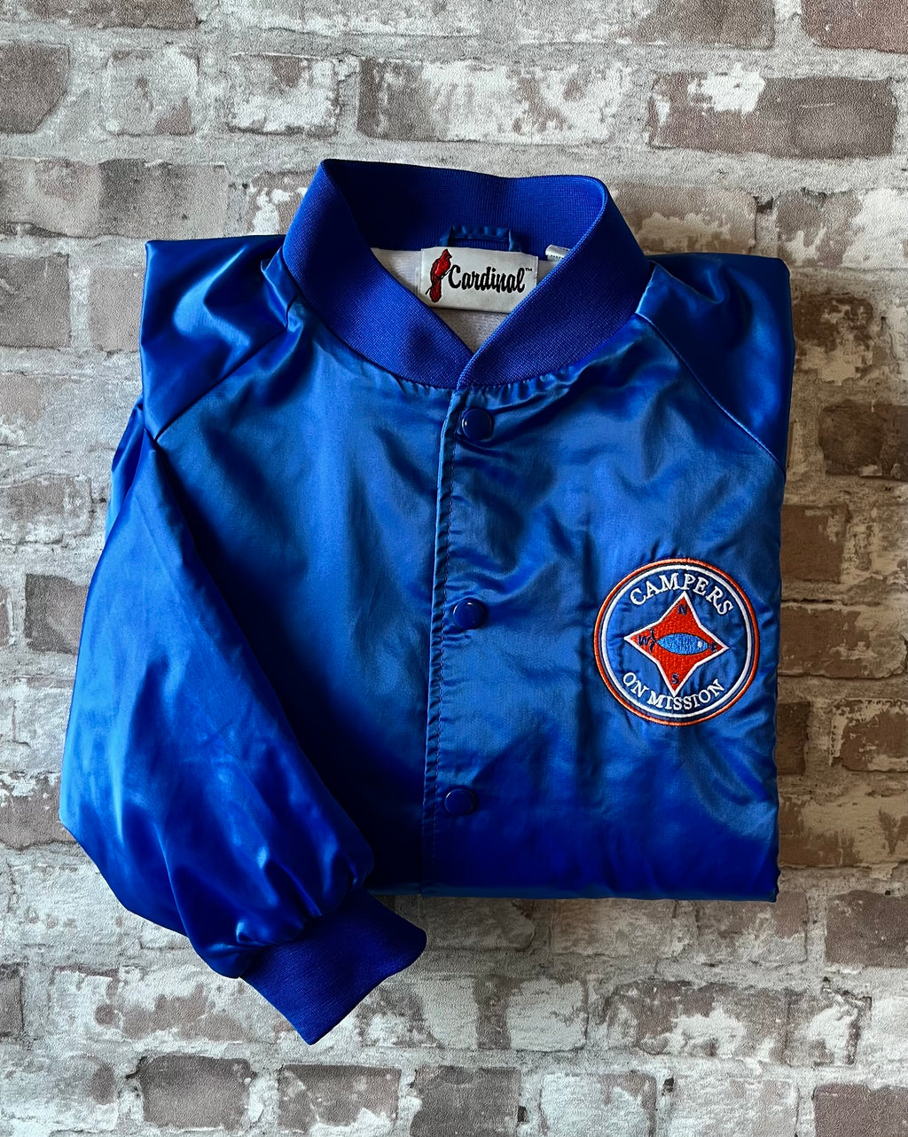 Veste de sport bomber léger rétro Cardinal bleu satiné avec fermeture à boutons-pression et écusson Campers On Mission, présentée à plat, pliée