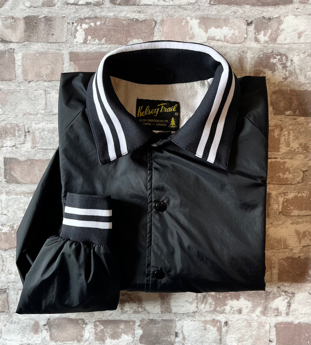 Veste coupe-vent vintage canadienne Kelsey Trail en nylon satiné noir avec boutons-pression et bords-côtes rayés, présentée pliée à plat.