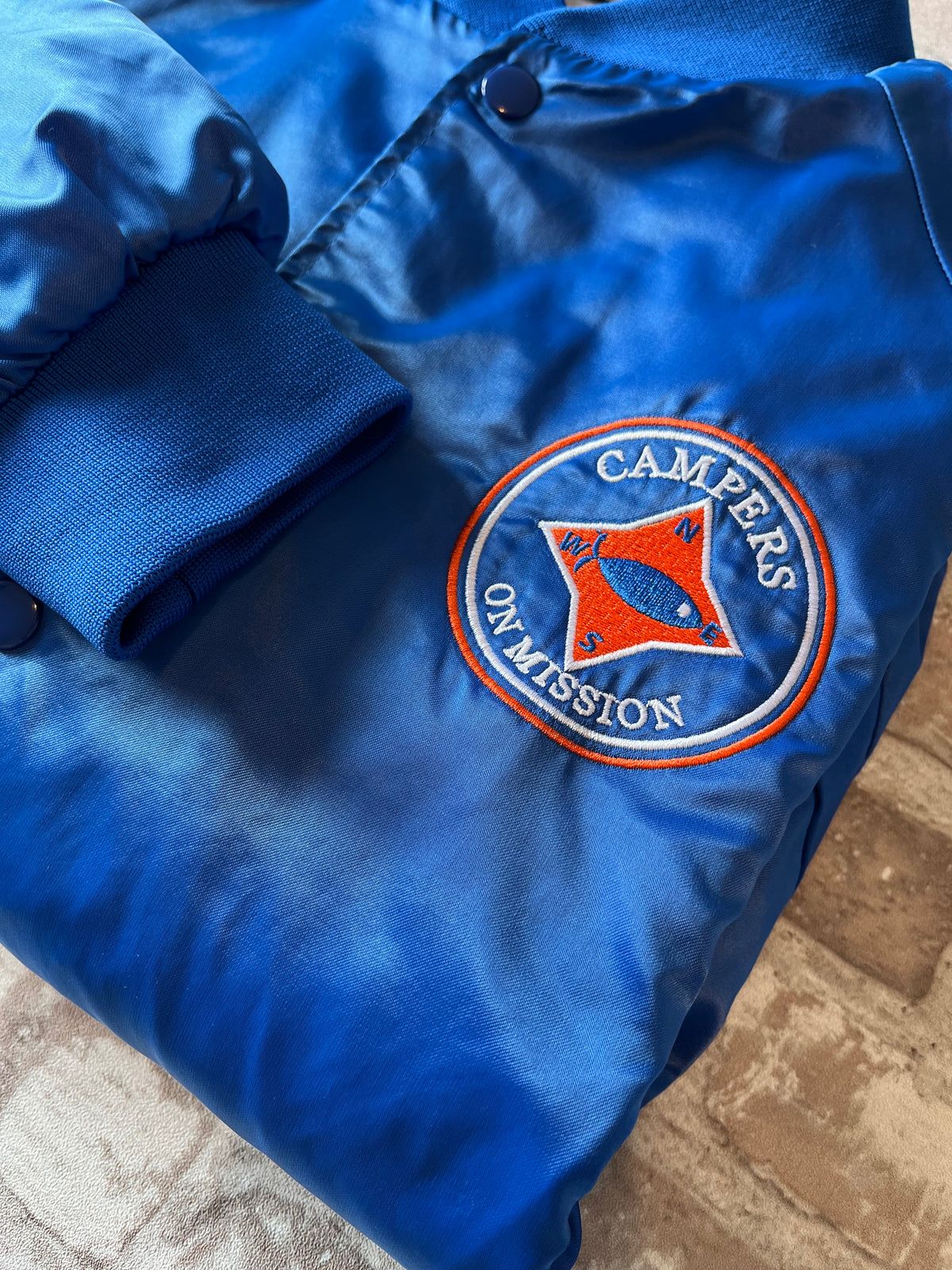 Veste de sport bomber léger rétro Cardinal bleu satiné avec fermeture à boutons-pression et écusson Campers On Mission, présentée à plat. Focus écusson