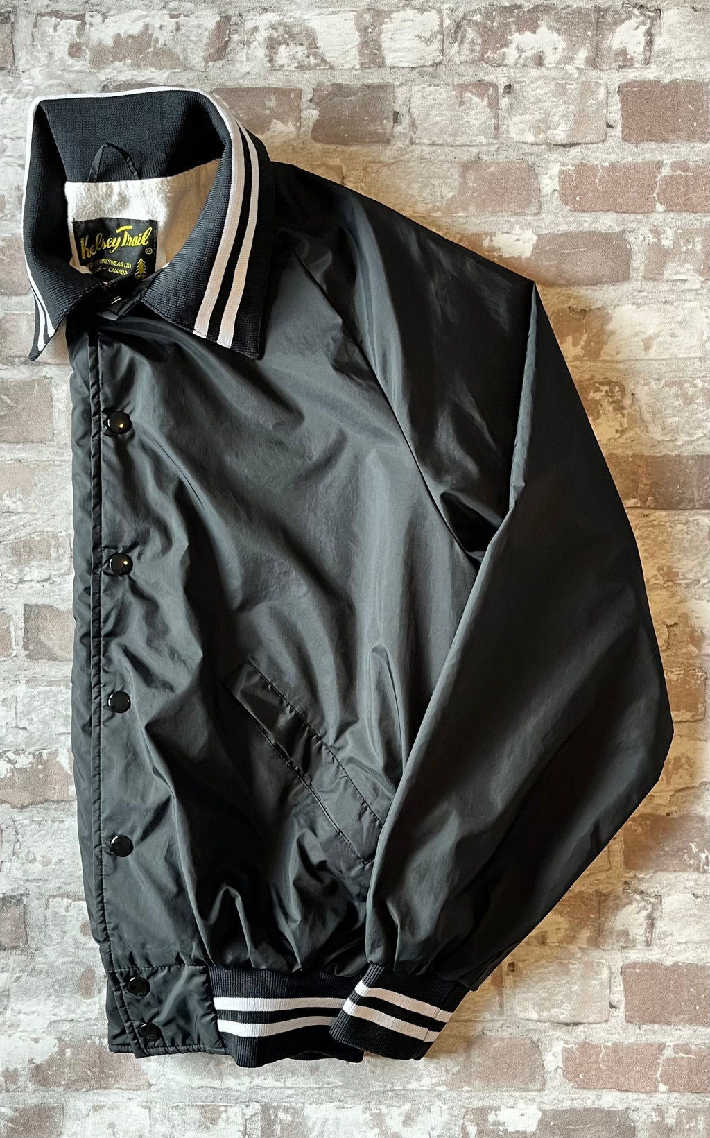 Veste coupe-vent vintage canadienne Kelsey Trail en nylon satiné noir avec boutons-pression et bords-côtes rayés, présentée pliée à plat de profil